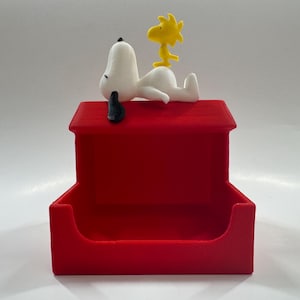 May include: A red, rectangular dog house-shaped container with a white Snoopy figure and a yellow Woodstock figure on top. The container has a small opening in the front.