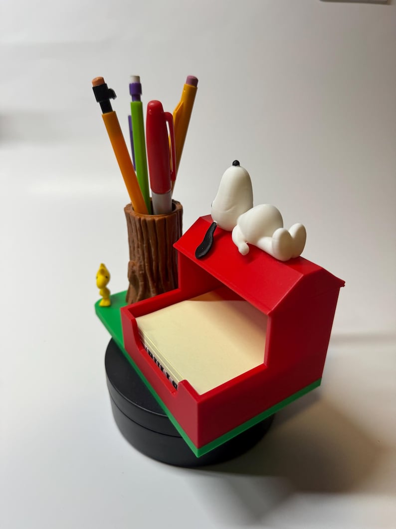 May include: A desk organizer featuring Snoopy on his red doghouse, with a brown tree trunk pen holder and a stack of notepaper. The organizer also includes a small yellow bird figure and several pens.