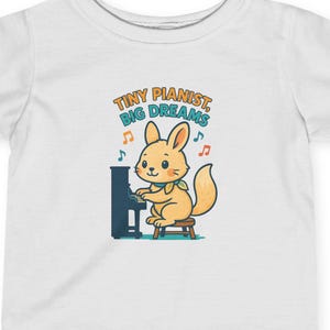 May include: White short-sleeved t-shirt featuring a cartoon rabbit playing a piano. The rabbit is light orange with a fluffy tail. The text "Tiny Pianist, Big Dreams" is above the rabbit in teal and orange.