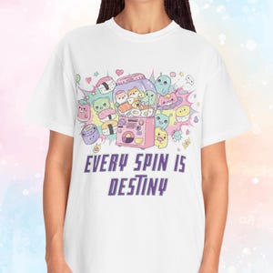 May include: White t-shirt featuring a colorful graphic of a gumball machine filled with cute cartoon characters, sushi, and other food items. The text "EVERY SPIN IS DESTINY" is printed below the graphic in purple.
