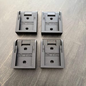 Könnte beinhalten: Vier schwarze DeWalt-Batteriehalter aus Kunststoff. Jeder Halter hat das DeWalt-Logo auf der Vorderseite aufgedruckt. Die Halter sind rechteckig mit einem Schlitz oben und einem kreisförmigen Loch in der Mitte.