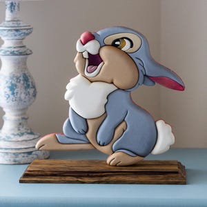May include: A wooden sculpture of a cartoon rabbit with blue, tan, white, and pink coloring. The rabbit is sitting on a wooden base. The rabbit has an open mouth and appears to be smiling. The sculpture is on a blue surface.