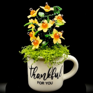 May include: A white ceramic mug with the black text thankful FOR YOU printed on the front. The mug is filled with a decorative arrangement of yellow glass flowers with orange centers and green glass leaves, set into a base of green moss.