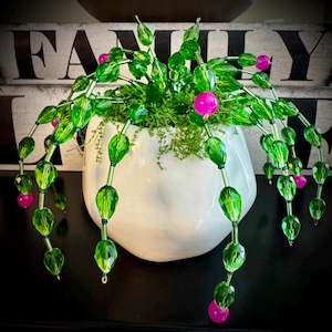 May include: A white ceramic pot filled with green moss and a beaded plant arrangement. The plant features green, faceted beads and pink spherical beads, with the word "FAMILY" visible in the background.