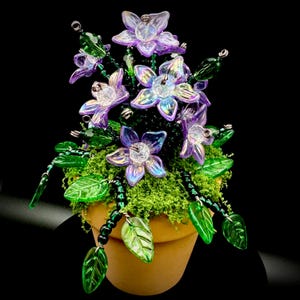 May include: A decorative arrangement featuring iridescent purple glass flowers with clear centers, accented by green glass leaves and dark green beaded stems, all set into a small terracotta pot filled with green moss on a black background.
