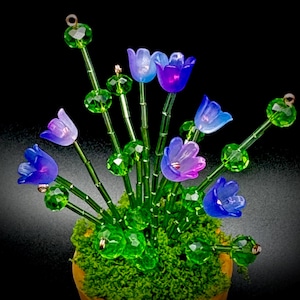 Könnte beinhalten: Ein handgefertigtes Blumenarrangement mit durchscheinenden blauen und lila tulpenförmigen Perlen. Die Stiele sind grün, und die Anordnung befindet sich in einem Bett aus grünem Moos. Die Perlen sind mit grünen, kristallartigen Perlen akzentuiert.