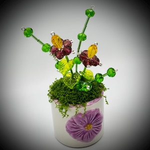 May include: A decorative floral arrangement in a white ceramic pot. The arrangement features beaded flowers in yellow and burgundy, with green leaves and stems. The pot is decorated with a purple flower design and filled with green moss.