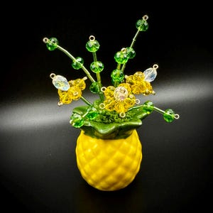 May include: A decorative pineapple-shaped vase in yellow ceramic, holding a beaded floral arrangement. The flowers are yellow and clear beads, with green beaded stems and leaves. The vase has a green rim and sits on a black surface.