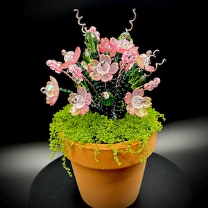 May include: A handcrafted floral arrangement in a terracotta pot. The arrangement features pink and clear beaded flowers, green beaded stems, and a bed of green moss. The pot sits on a black surface.