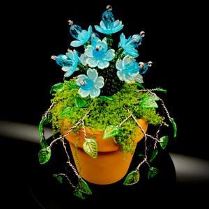 May include: A decorative potted plant sculpture featuring light blue translucent glass flowers with gold-toned edges, green glass leaves on twisted wire stems, and a base of green moss inside a small, round, terracotta-colored clay pot.