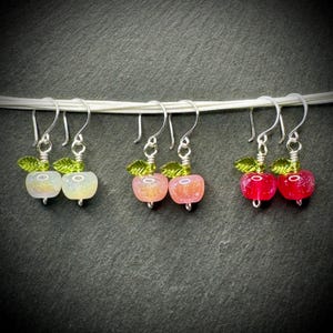 Puede incluir: Tres pares de pendientes con forma de manzana. Cada pendiente presenta una manzana de cristal en tonos blanco, rosa y rojo, con detalles de hojas verdes y ganchos plateados. Los pendientes se muestran sobre una cuerda blanca sobre un fondo oscuro.