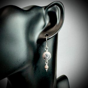 May include: A single dangle earring with a silver hook and wirework. The earring features a large, round, pink and gray stone, with a smaller, round, pink stone below. The earring is displayed on a black mannequin head.