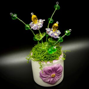 May include: A decorative floral arrangement in a white ceramic pot. The arrangement features beaded flowers with purple and yellow accents, green stems, and a bed of green moss. A purple flower is embossed on the pot.
