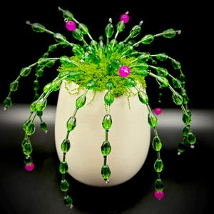 May include: A decorative beaded plant arrangement in a cream-colored pot. The plant features green beaded stems with pink accents and is set against a black background. The arrangement includes a bed of green moss.