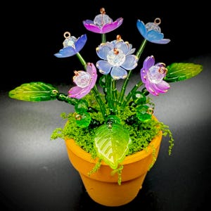 Puede incluir: Un arreglo floral en miniatura en una maceta de terracota. Presenta delicadas flores translúcidas en tonos azules, morados y blancos, con detalles dorados. Hojas verdes y musgo completan el diseño, sobre un fondo negro.