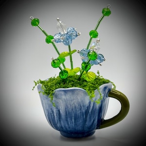 May include: A decorative floral arrangement in a blue teacup. The arrangement features beaded flowers in shades of green, blue, and clear, set in a bed of green moss. The teacup has a green handle and a textured blue exterior.