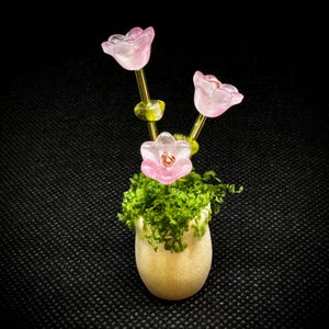 May include: A miniature floral arrangement featuring three pink tulip-shaped flowers with gold stems and green accents, emerging from a small, off-white pot filled with green moss. The background is black.