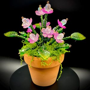 Op de afbeelding: Een handgemaakt bloemstuk in een terracotta pot. Het arrangement heeft roze, doorschijnende bloemen met gouden accenten, groene stelen en bladeren. De pot is gevuld met groen mos. De achtergrond is zwart.