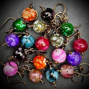 May include: A collection of handmade earrings featuring colorful, marbled glass beads in various hues including red, green, purple, and orange. Each bead is suspended from a gold-toned wire hook, creating a unique and artistic jewelry design.