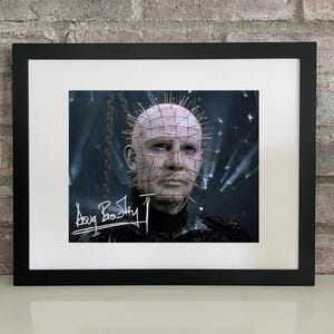 May include: Framed print featuring a character with pins in their head, a signature, and a black frame. The image has a dark, cinematic aesthetic. The signature reads "Doug Bradley".
