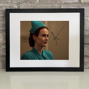 May include: Framed print featuring a woman in a teal uniform and hat, with a signature in black ink. The image is set against a neutral background and framed in a black border. The woman has brown hair and is wearing earrings.
