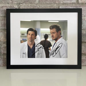 Puede incluir: Fotografía enmarcada con dos hombres con batas blancas, probablemente médicos, con firmas. La imagen está ambientada sobre un fondo borroso de hospital. El marco es negro y el fondo es una pared de ladrillos.