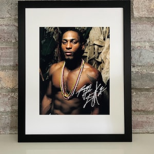 Puede incluir: Impresión enmarcada de un hombre con collares, en un marco negro con un paspartú blanco. La imagen muestra a un hombre con un tatuaje y una firma. El fondo presenta tonos verdes y marrones. La impresión se muestra sobre una superficie blanca.