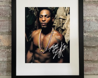 May include: Framed print featuring a shirtless man wearing beaded necklaces. The print is in a black frame with a white mat. The man has a tattoo on his arm and a signature in the bottom right corner. The background is a mix of green and brown.