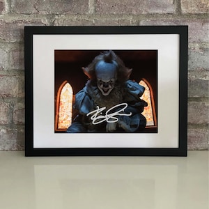 May include: Framed print featuring a character from a horror film. The image shows a menacing figure in a clown-like costume, with a white face and red hair. The print is signed in white ink and set against a background of stained-glass windows.