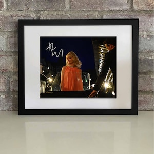 May include: Framed photograph featuring a woman in an orange dress, with an autograph in the upper left corner. The image is set against a night scene with lights. The frame is black, and the photograph is mounted on a white mat.