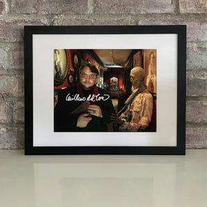 Puede incluir: Impresión enmarcada en negro con paspartú blanco, que muestra a Guillermo del Toro sosteniendo un libro, firmado en blanco. El fondo muestra una habitación con retratos y una figura.