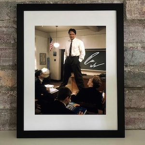 May include: Framed print depicting a scene from a film. A man in a white shirt and black trousers stands on a desk in a classroom. The image includes a blackboard with a signature and students in the background.