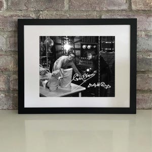 Op de afbeelding: Zwart omlijste zwart-witfoto met een scène uit een klassieke film. De afbeelding toont twee mannen in een laboratorium, met handtekeningen rechtsonder. De foto is voorzien van een witte rand.