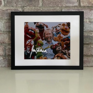 Puede incluir: Fotografía enmarcada con Jim Henson y personajes de los Muppets, firmada con tinta blanca. El marco negro tiene un paspartú blanco, sobre un fondo de pared de ladrillo.