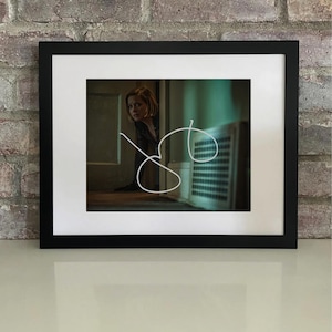 Puede incluir: Impresión artística enmarcada que muestra a una mujer mirando por una puerta. La imagen tiene una estética oscura y melancólica, con un marco negro y un paspartú blanco. Un dibujo lineal blanco se superpone a la imagen. El fondo es una pared de ladrillos.