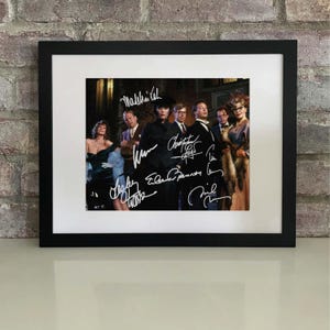 May include: Black framed photograph with a white mount, featuring a group of people, possibly actors, with signatures. The background is a brick wall. The image is a portrait.
