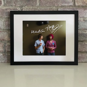 May include: Framed photograph featuring two people standing in front of a wall. The photograph is signed with the names "Michael Cera" and "Mary Elizabeth Winstead". The people are holding red cups. The frame is black with a white mat.