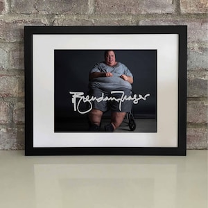 May include: Framed print featuring a portrait of a person in a wheelchair, wearing a gray t-shirt and shorts. The print is signed with the name "Brendan Fraser" in white script. The frame is black, and the background is a dark gray.