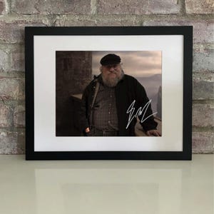 Może przedstawiać: Oprawiona fotografia przedstawiająca mężczyznę z długą białą brodą, ubranego w czarną kurtkę i czapkę. Fotografia podpisana białym tuszem. Rama jest czarna, a fotografia umieszczona na białym passe-partout.