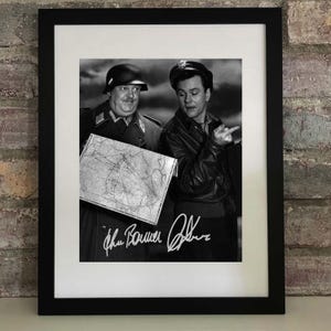 May include: Black and white framed print featuring two men in military-style attire. One man holds a map, while the other points. The print is signed at the bottom. The frame is black.