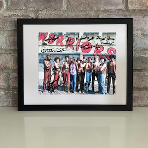 Peut inclure: Tirage encadré noir représentant un groupe de personnes posant devant un graffiti rouge "WARRIORS". L'impression est signée par plusieurs personnes.