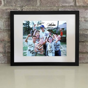 May include: Framed photograph featuring a group of children, likely from a movie or show, with signatures. The photo is in a black frame with a white mat. The children are wearing baseball uniforms and casual clothes, celebrating with raised fists.