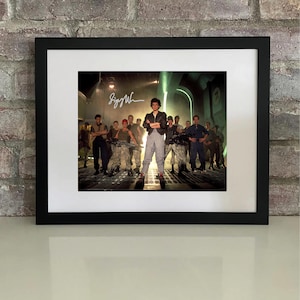 May include: Framed print featuring a movie scene with a central figure in a black jacket and gray pants, standing confidently. The image is signed and surrounded by a group of people in military-style attire. The print is set in a black frame.