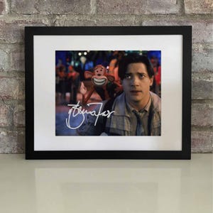 May include: Framed print featuring a movie still with a man and a puppet. The image is signed in white ink. The frame is black, and the print is set against a white mat. The background is a brick wall.