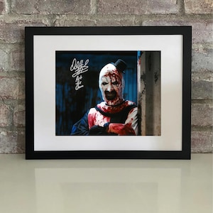 May include: Framed print featuring a character in clown makeup with blood-like stains. The clown wears a small top hat and a dark outfit. The print is in a black frame with a white mat, displayed against a brick wall.