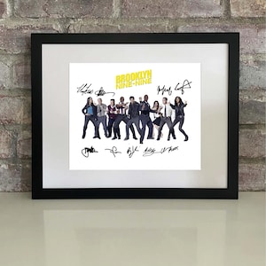 May include: Framed print featuring the cast of Brooklyn Nine-Nine, with the show's title in yellow. The image is surrounded by a black frame and includes signatures of the cast members. The print is displayed against a brick wall.