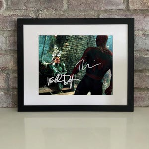 May include: Framed print depicting a movie scene with two costumed figures. The print is signed in white ink. The frame is black, and the print has a white border. The background is a brick wall.