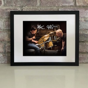 May include: Framed photograph featuring a drummer playing a drum kit, with a second person observing. The image is signed "Matt Oasis" in white ink. The photograph is set in a black frame with a white mat, against a brick wall.