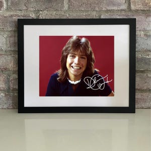 Può includere: Fotografia incorniciata con una persona sorridente con lunghi capelli castani, che indossa una camicia blu scuro e una collana bianca. Lo sfondo è rosso pieno. La fotografia è firmata con una firma bianca.