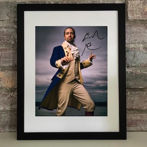 May include: Framed photograph of a man in a historical costume, likely from a stage production. He wears a blue coat with gold trim, a cream-colored vest, and tan pants. The photo is signed in black ink.